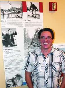Dion Agee standing for a photo at Mammoth Lakes Foundation offices next to a display featuring his grandfather, Bob Bumbaugh, and Dave McCoy. Photo courtesy: Sheet staff.